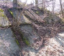 Uferdeckwerk aus Platten zum Schutz vor Seitenerosion. Die Platten sind stark von Vegetation überwachsen und teilweise verschoben und abgesackt. Das Schutzbauwerk ist in einem schlechten Zustand.